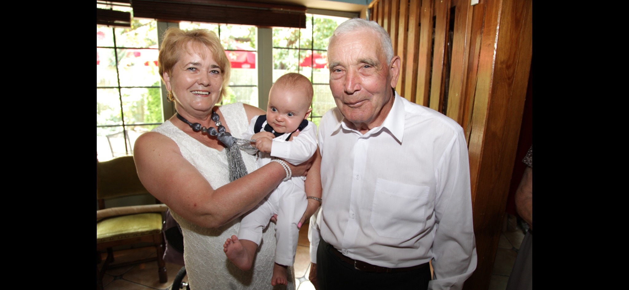 Uśmiechnięta kobieta w białej sukience i z naszyjnikiem trzyma na rękach niemowlę ubrane w biały kombinezon z granatową muszką, obok stoi starszy mężczyzna w białej koszuli, w tle okno z zielenią.
