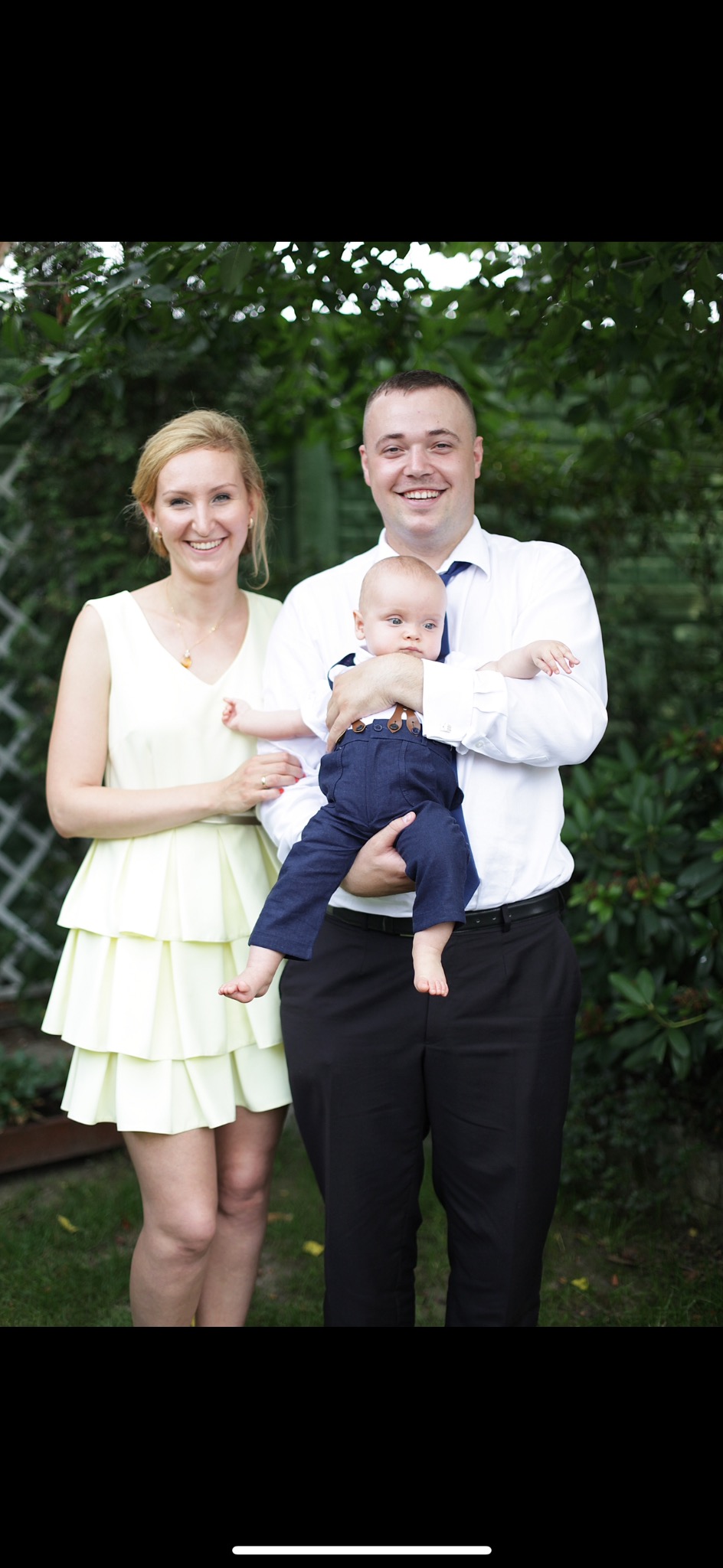 Uśmiechnięta rodzina: mama w żółtej sukience, tata trzymający niemowlę w granatowym ubranku z szelkami, na tle zieleni.