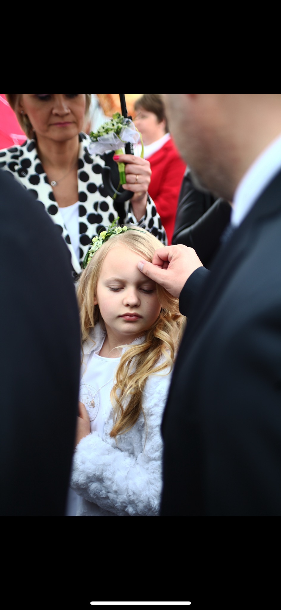 Dziewczynka z blond włosami i wiankiem na głowie, w białym ubranku, podczas błogosławieństwa, z zamkniętymi oczami, dotykana przez dłoń mężczyzny w garniturze.