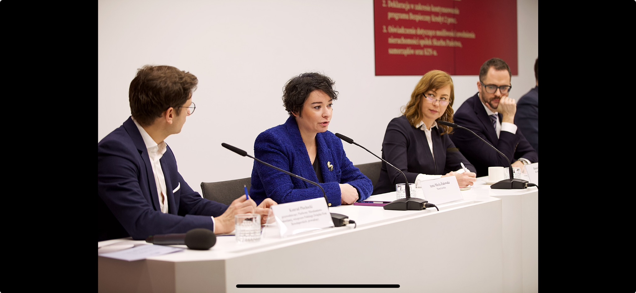 Kobieta w niebieskiej marynarce przemawia podczas konferencji, widoczni inni uczestnicy, mikrofony i tabliczki z nazwiskami.