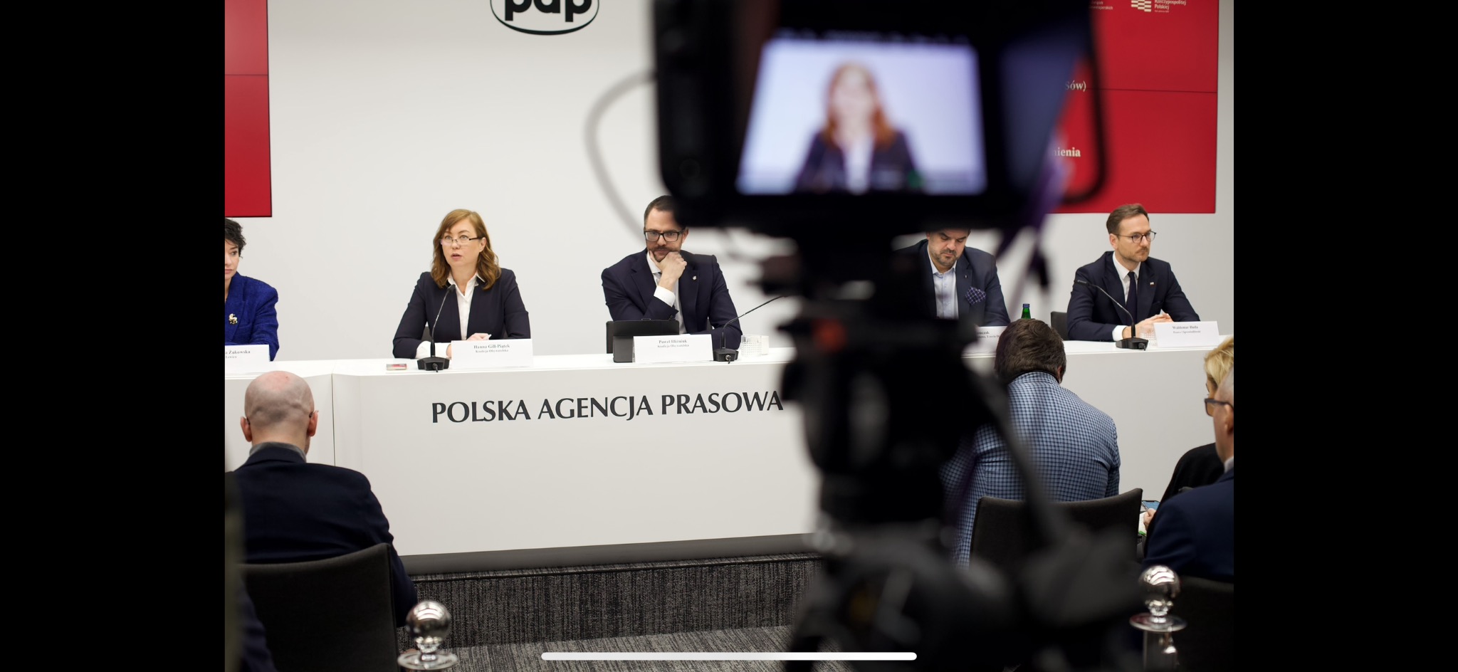 Konferencja prasowa Polskiej Agencji Prasowej, widok przez obiektyw kamery, prezenterzy siedzący za stołem, publiczność na pierwszym planie.