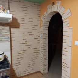 Kuchnia po remoncie z dekoracyjną ścianą z imitacji kamienia i łukiem nad przejściem do innego pomieszczenia. Zielony sufit i pomarańczowa ściana. Widoczny fragment kuchenki i dekoracje na szafce.