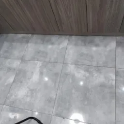 Świeżo wykafelkowana podłoga w łazience z szarymi, matowymi płytkami imitującymi kamień, zestawiona z panelami ściennymi imitującymi drewno, widoczne fragmenty czarnego przedmiotu w rogu kadru.