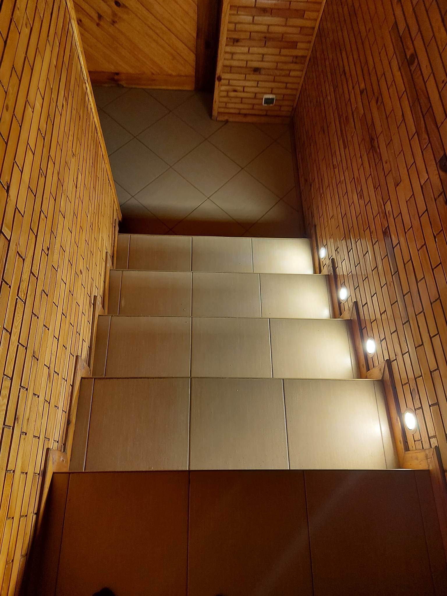 Wykończone schody z jasnymi płytkami, drewnianą boazerią w stylu cegły i punktowym oświetleniem LED na ścianie.