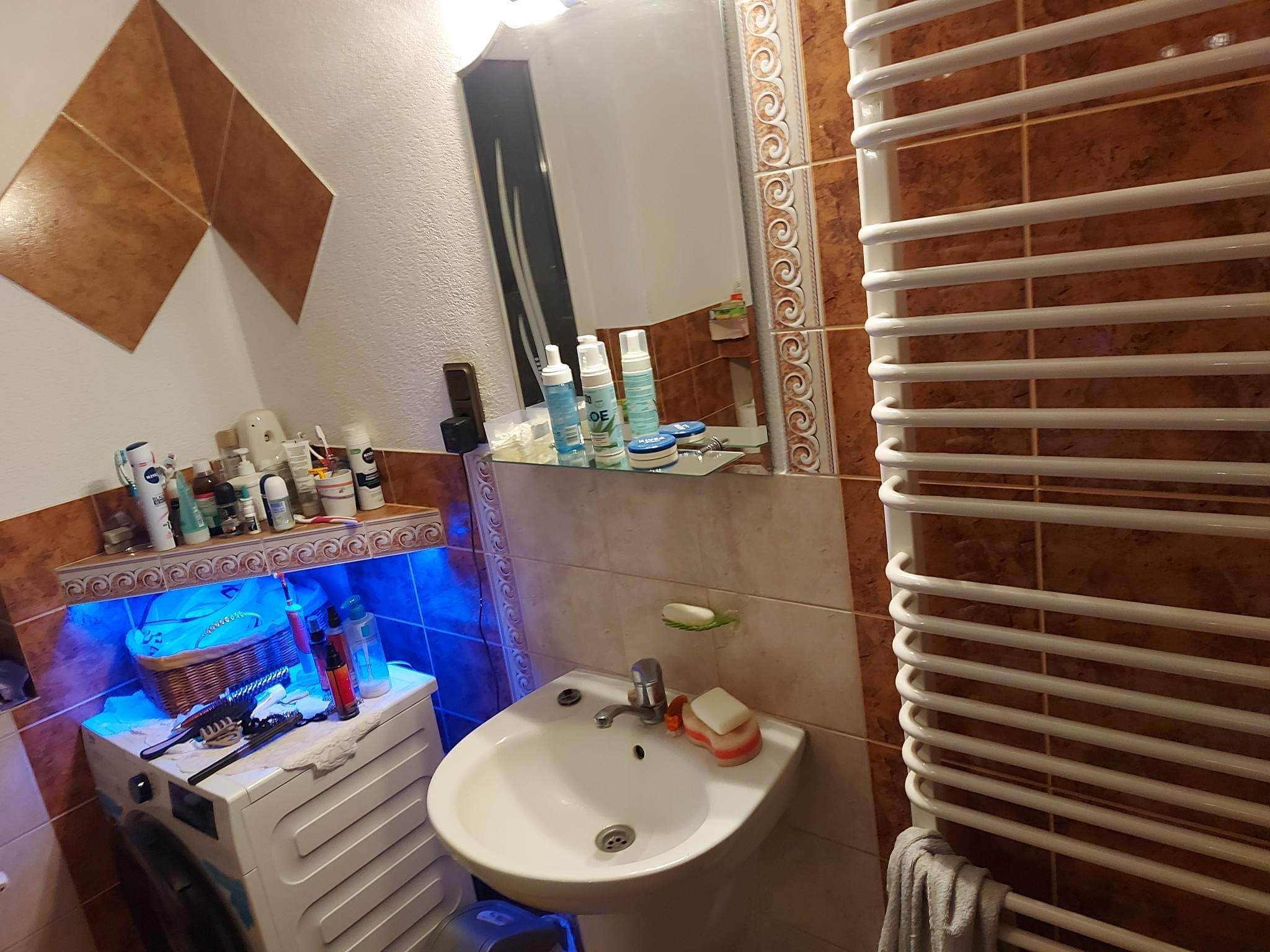 Umywalka z baterią i mydłem w łazience z brązowymi płytkami, obok pralka z koszem i niebieskim podświetleniem półki, na ścianie grzejnik drabinkowy