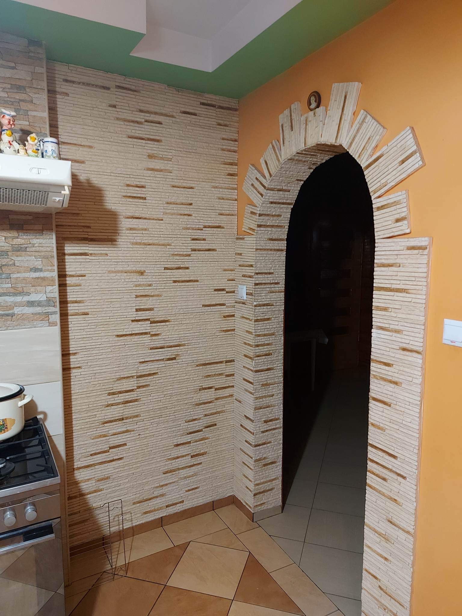 Kuchnia po remoncie z dekoracyjną ścianą z imitacji kamienia i łukiem nad przejściem do innego pomieszczenia. Zielony sufit i pomarańczowa ściana. Widoczny fragment kuchenki i dekoracje na szafce.