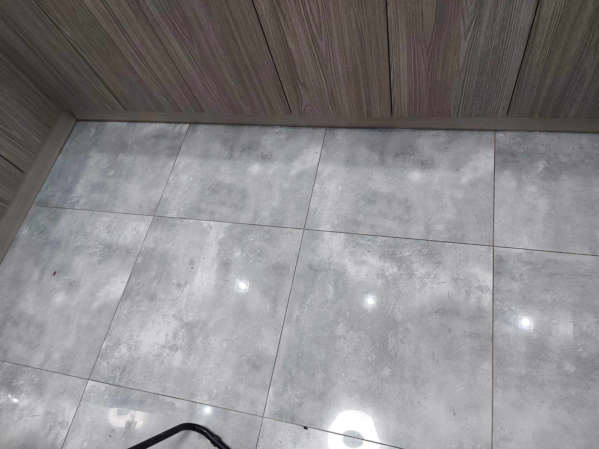 Świeżo wykafelkowana podłoga w łazience z szarymi, matowymi płytkami imitującymi kamień, zestawiona z panelami ściennymi imitującymi drewno, widoczne fragmenty czarnego przedmiotu w rogu kadru.