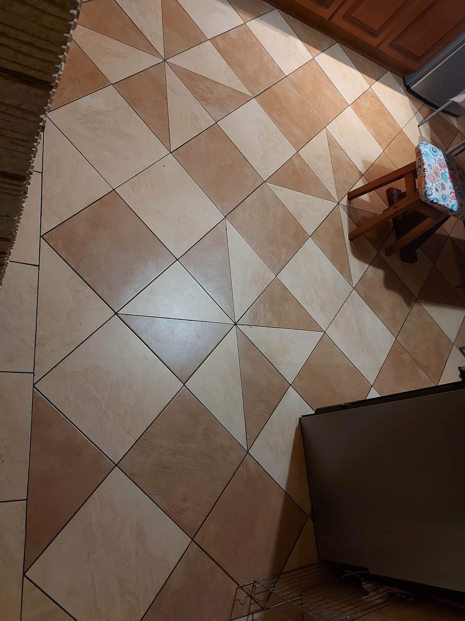 Ułożona pod kątem podłoga z kwadratowych, beżowo-brązowych płytek ceramicznych, fragment szafki kuchennej, krzesło z kwiecistym siedziskiem.