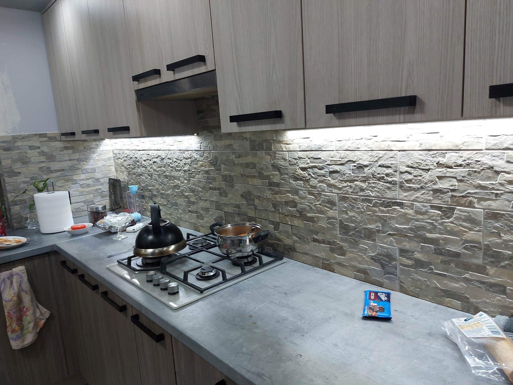 Nowoczesna kuchnia z szarymi szafkami, czarnymi uchwytami i kamienną mozaiką na ścianie, oświetlona listwą LED pod szafkami, z czajnikiem i garnkiem na płycie gazowej.