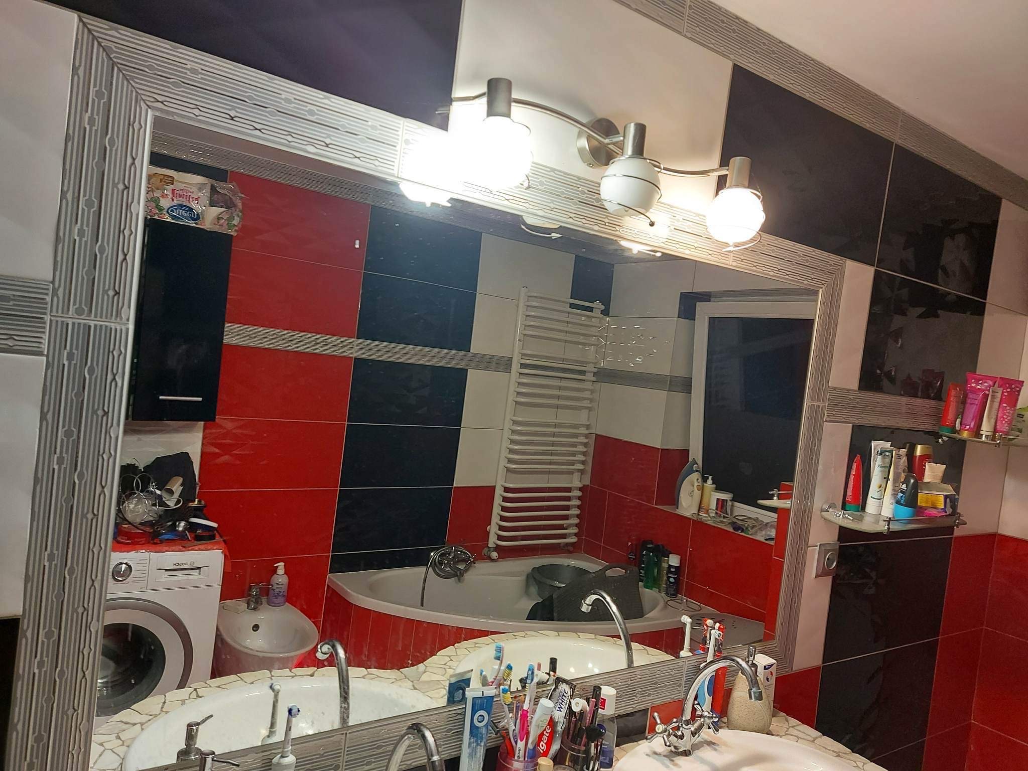 Wnętrze łazienki z umywalkami odbite w dużym lustrze, widoczna pralka, wanna narożna, grzejnik drabinkowy i płytki w kolorze czerwonym, czarnym i białym.