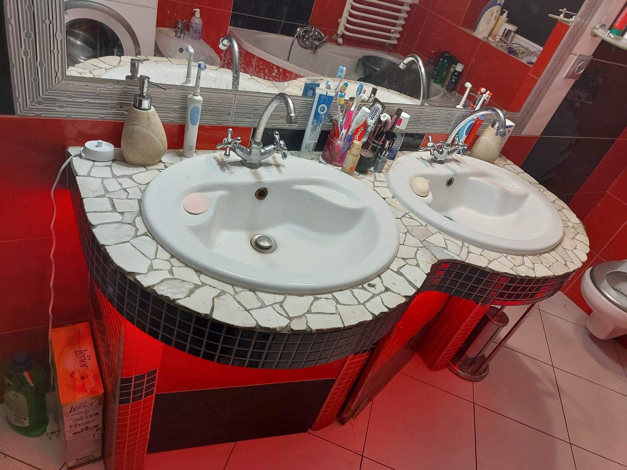 Dwie białe umywalki z bateriami, otoczone mozaiką z białych i czarnych płytek, w łazience z czerwonymi ścianami i płytkami podłogowymi, widoczne akcesoria łazienkowe i pralka w tle.