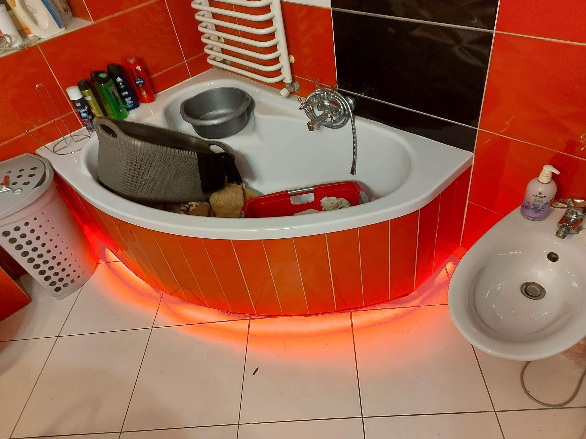 Wanna narożna z podświetleniem LED w łazience z pomarańczowo-czarnymi płytkami, widoczna umywalka, grzejnik drabinkowy i akcesoria łazienkowe.