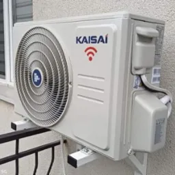 Zewnętrzna jednostka klimatyzatora Kaisai zamontowana na ścianie budynku, widoczna z boku, z fragmentem balkonu i okna w tle.
