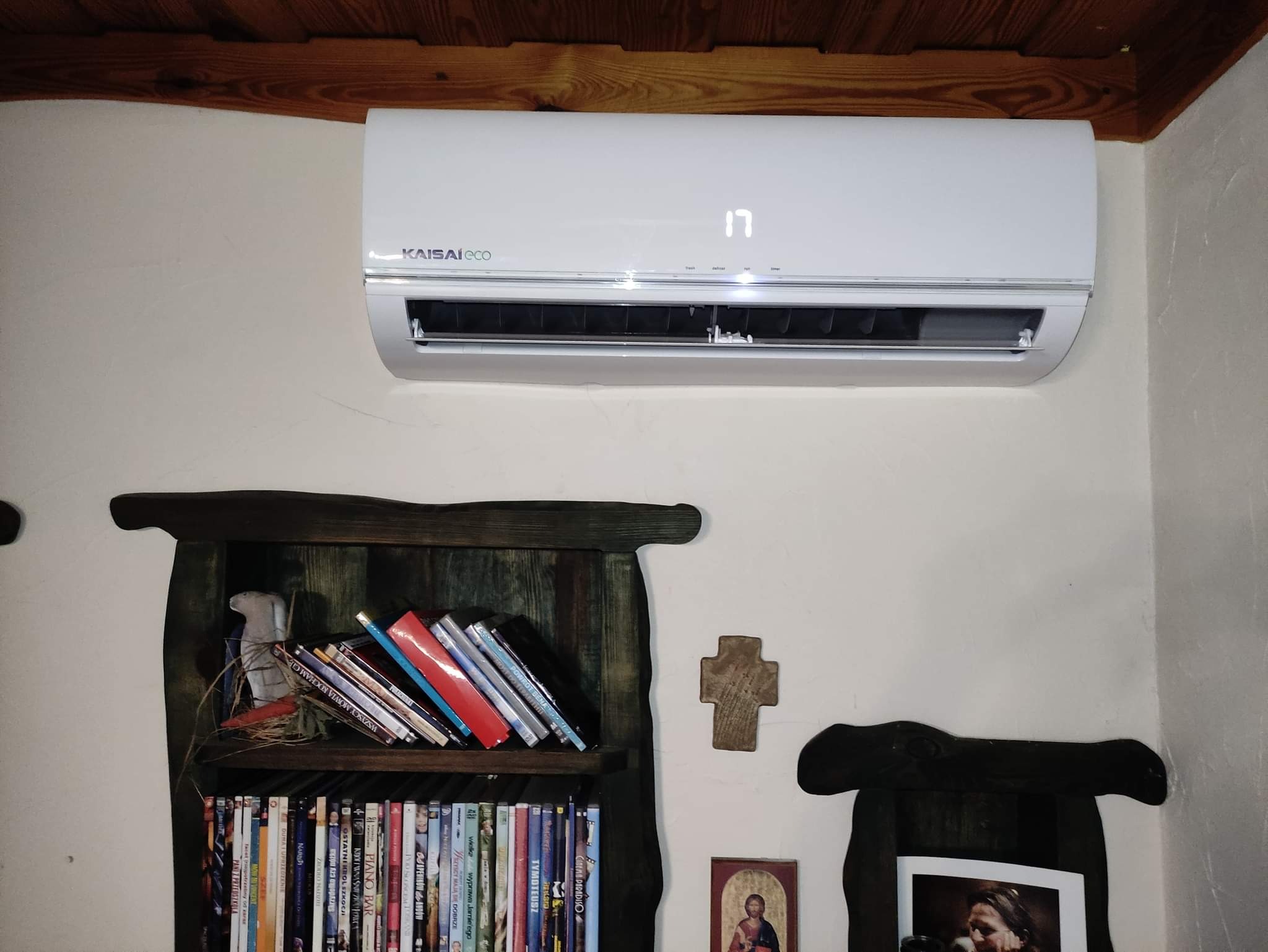 Biały klimatyzator Kaisai Eco z wyświetlaczem temperatury '17' zamontowany na ścianie nad półką z książkami i filmami DVD w rustykalnym wnętrzu z drewnianym sufitem.
