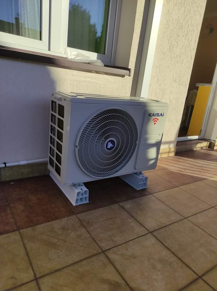 Zewnętrzna jednostka klimatyzacji Kaisai, zamontowana na białych wspornikach, na tle beżowej elewacji budynku w Wyszkowie, z widocznym oknem i brązowym parapetem.