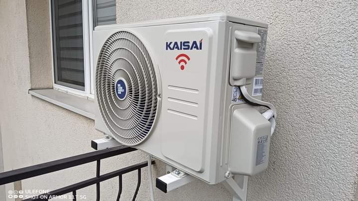 Zewnętrzna jednostka klimatyzatora Kaisai zamontowana na ścianie budynku, widoczna z boku, z fragmentem balkonu i okna w tle.