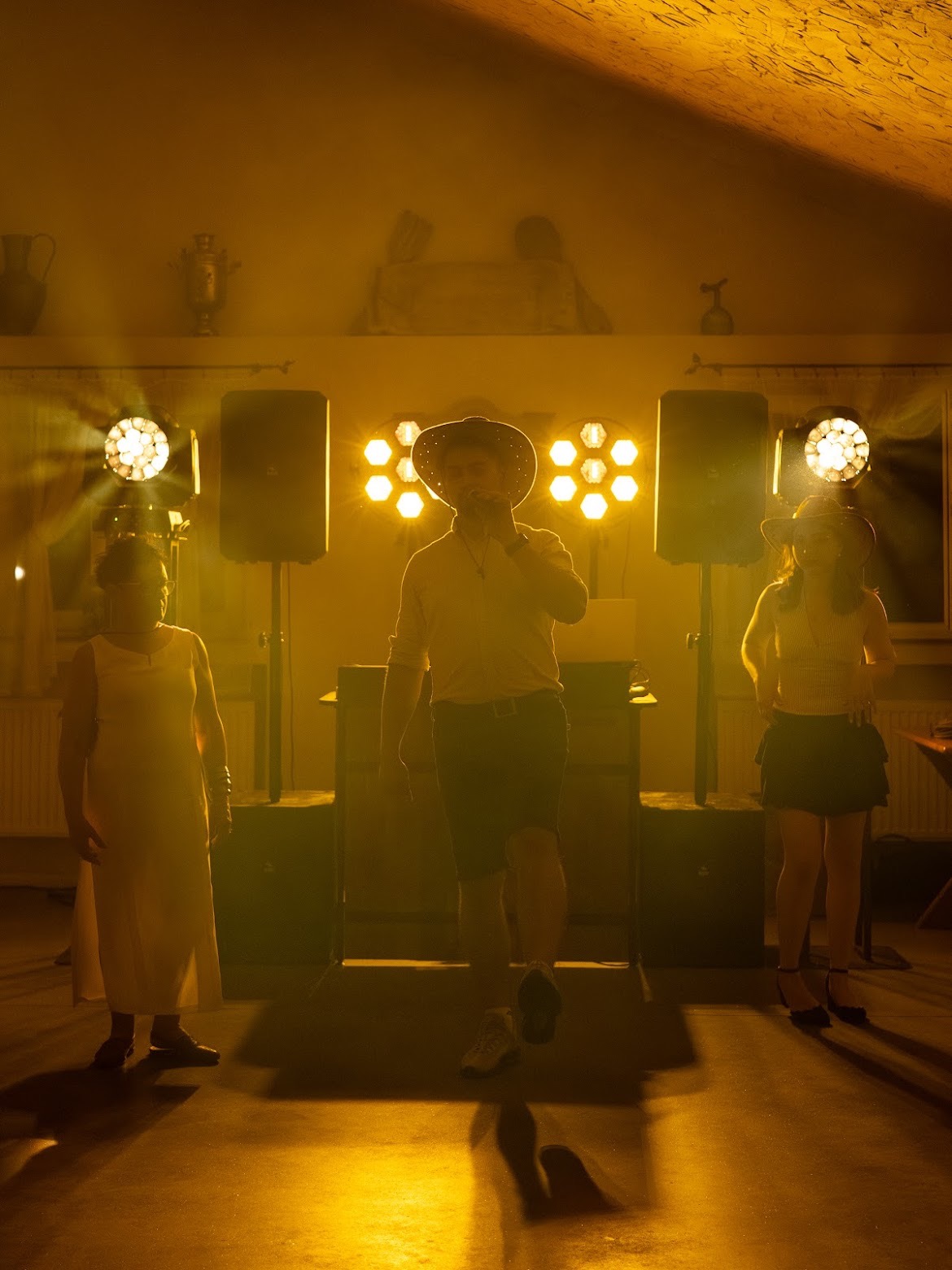 Zespół country na scenie z oświetleniem: wokalista w kapeluszu i dwie kobiety w kapeluszach. Scena oświetlona ciepłym, żółtym światłem. Dekoracje w tle.