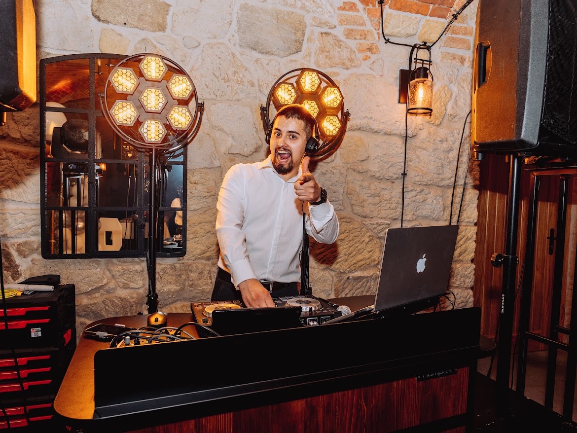 Uśmiechnięty DJ z brodą, w słuchawkach, miksuje muzykę na tle kamiennej ściany z dekoracyjnym oświetleniem i sprzętem nagłośnieniowym. Radosny gest dłoni.