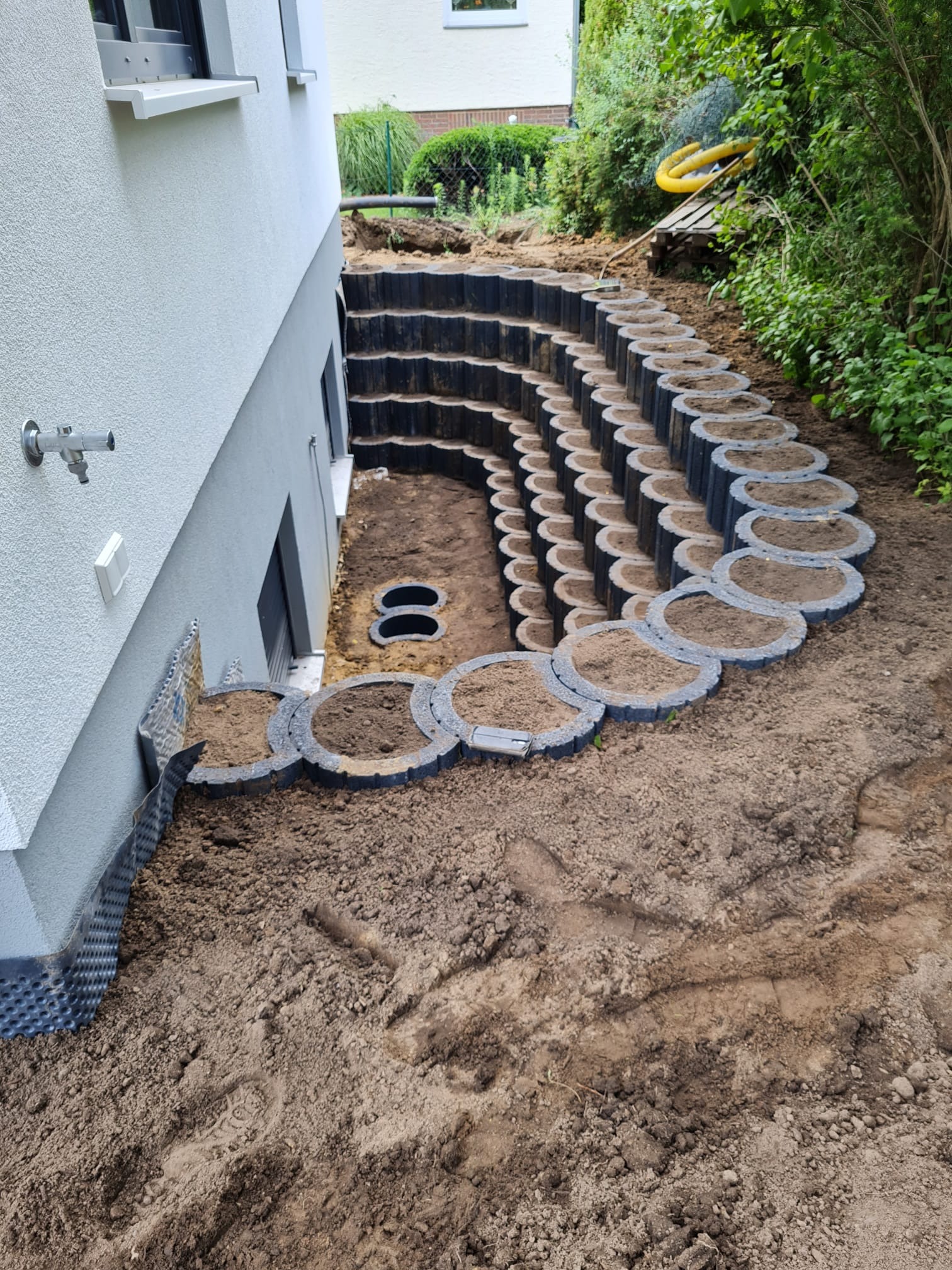 Spiralne schody z prefabrykowanych elementów betonowych, wypełnione ziemią, prowadzące do piwnicy budynku. Widoczne ślady stóp na ziemi i fragment izolacji fundamentów przy ścianie budynku.