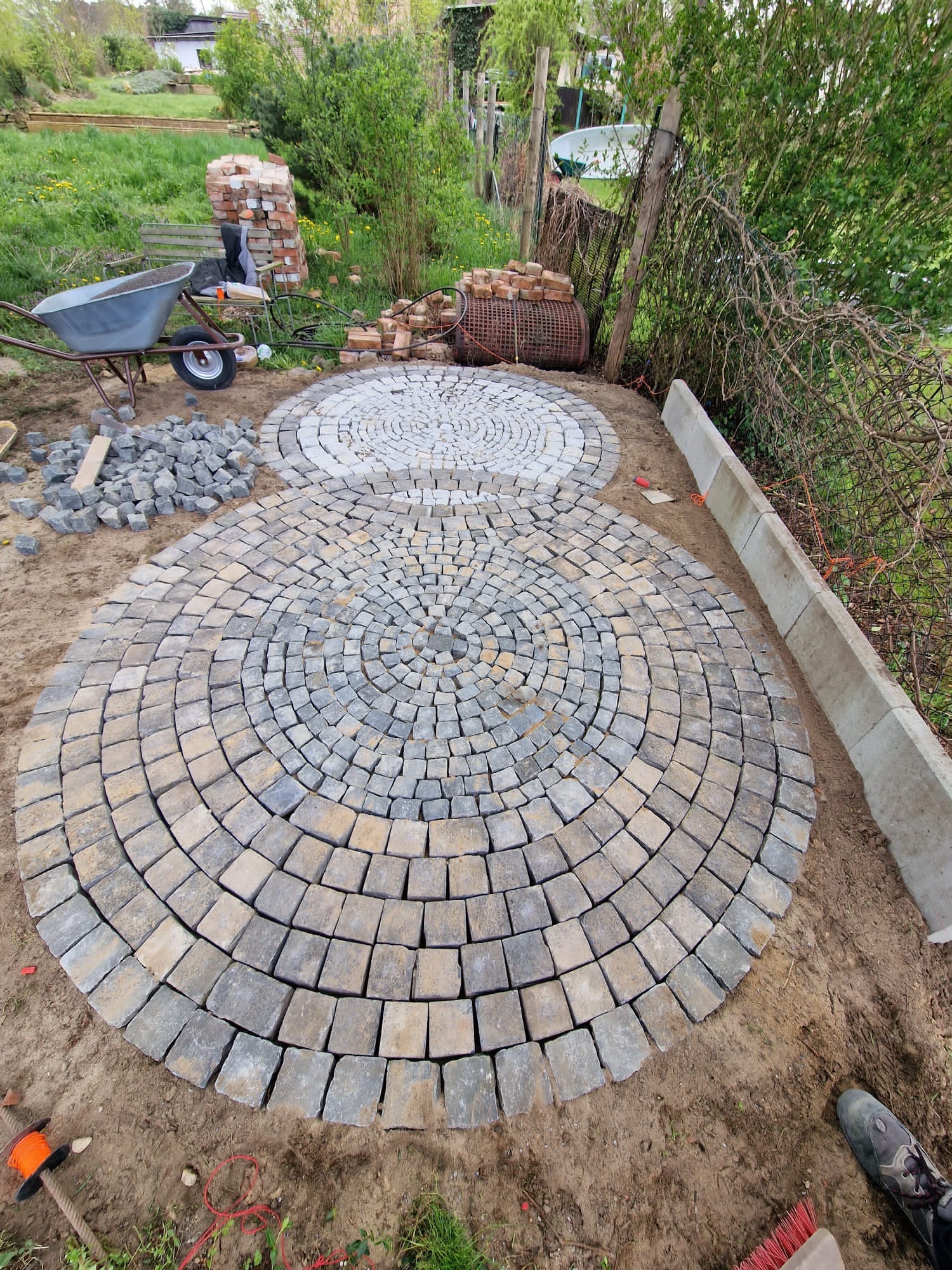 Dwa okręgi wyłożone kostką brukową w odcieniach szarości i beżu, ułożone koncentrycznie, otoczone ziemią i fragmentem trawnika, widoczna taczka z materiałami budowlanymi w tle.