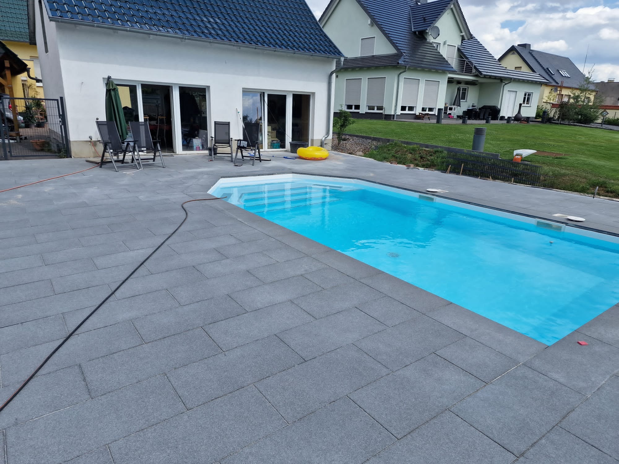 Nowoczesny basen otoczony szarą kostką brukową, widok z perspektywy ziemi, w tle biały dom z ciemnym dachem i zadbany trawnik.