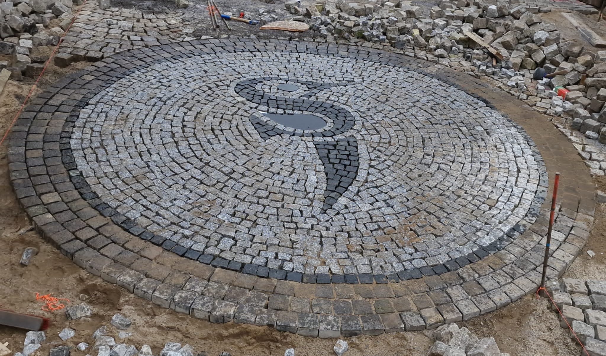 Okrągła powierzchnia wyłożona kostką brukową w odcieniach szarości, z ciemnoszarym wzorem przypominającym literę S, otoczona ciemniejszym obramowaniem z kostki i luźno ułożonymi kamieniami w tle.