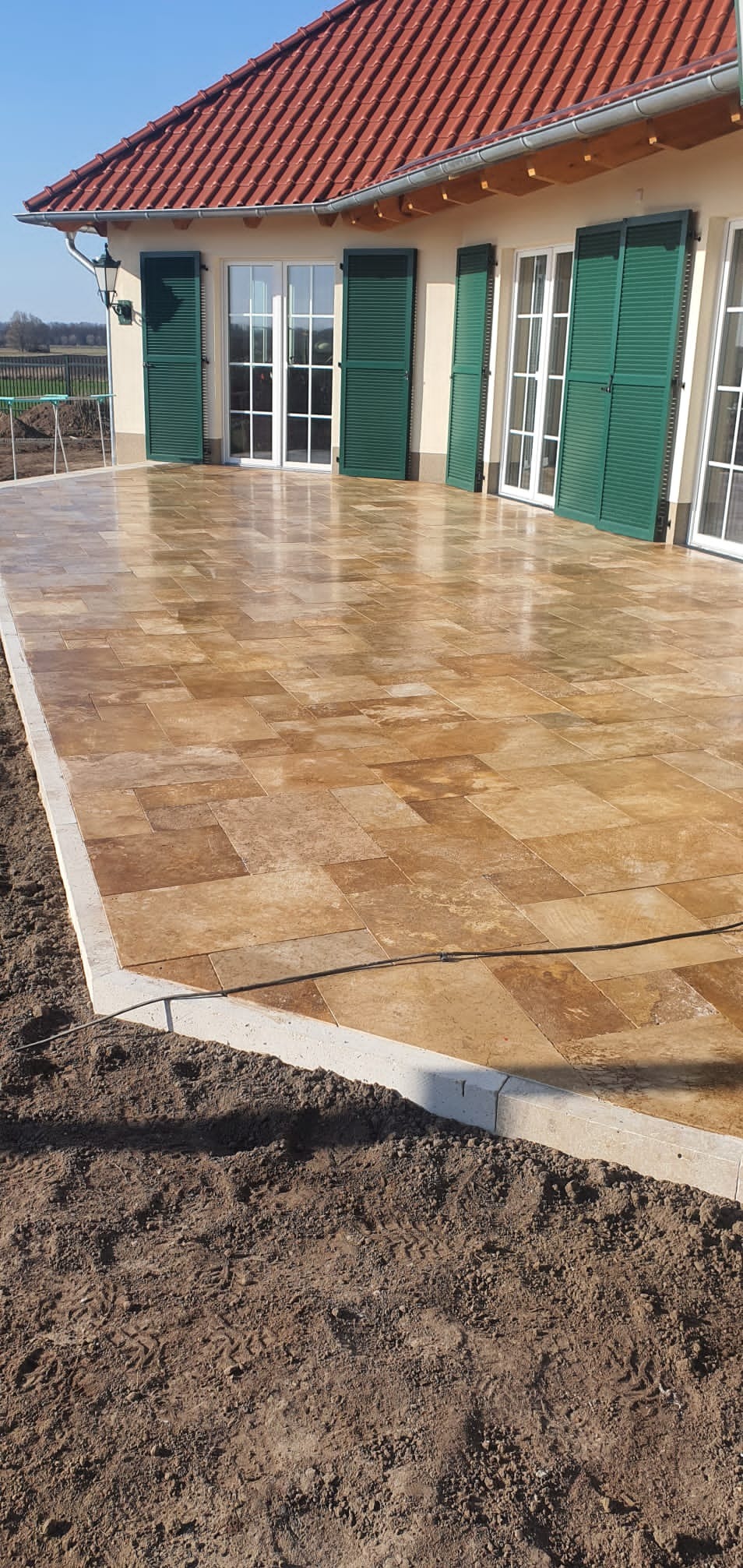 Nowo ułożona, brązowa kostka brukowa na tarasie domu z zielonymi okiennicami i czerwoną dachówką, otoczona świeżo przekopaną ziemią.