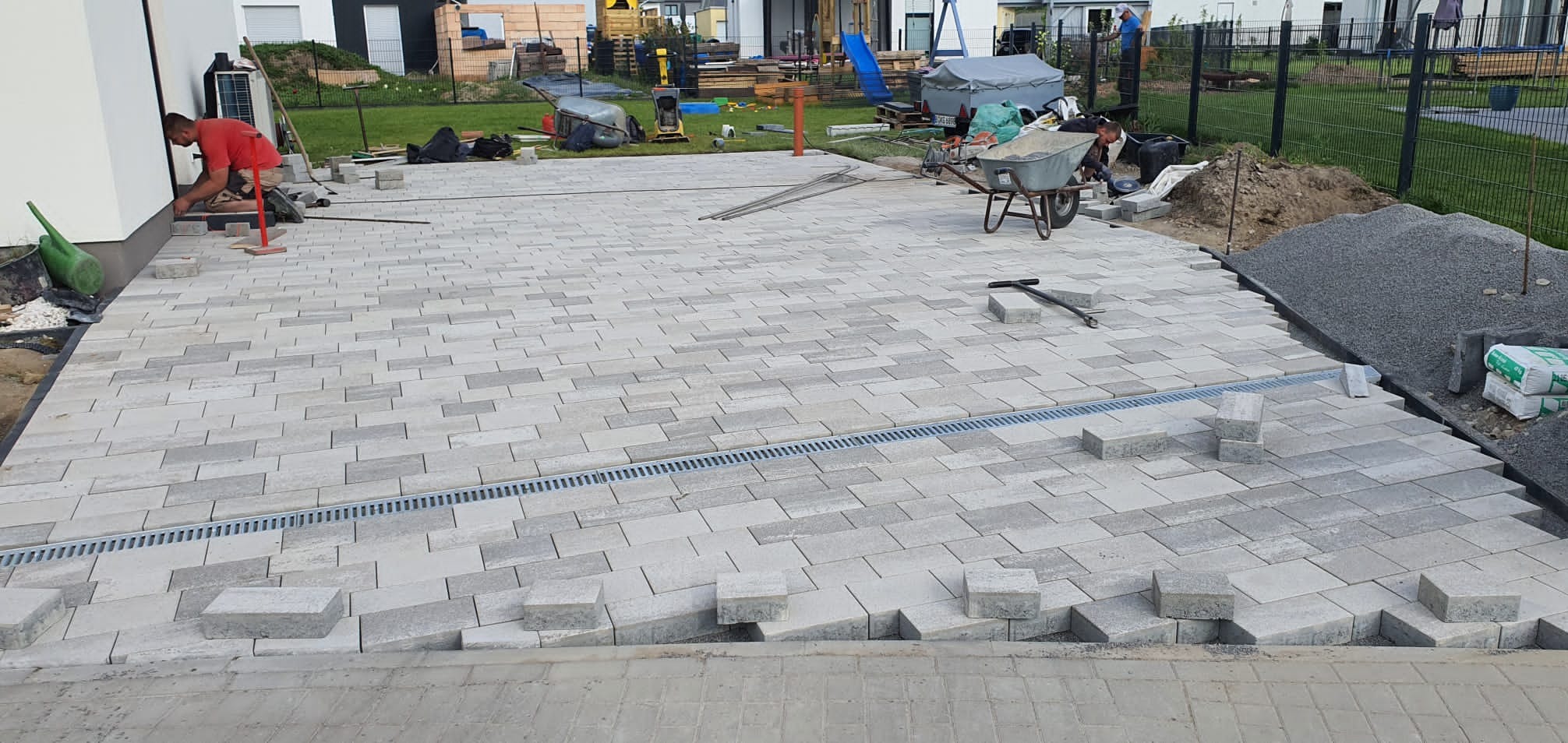 Układanie szarej kostki brukowej na posesji z widocznym drenażem liniowym, pracownikiem w czerwonej koszulce, narzędziami i materiałami budowlanymi w tle.