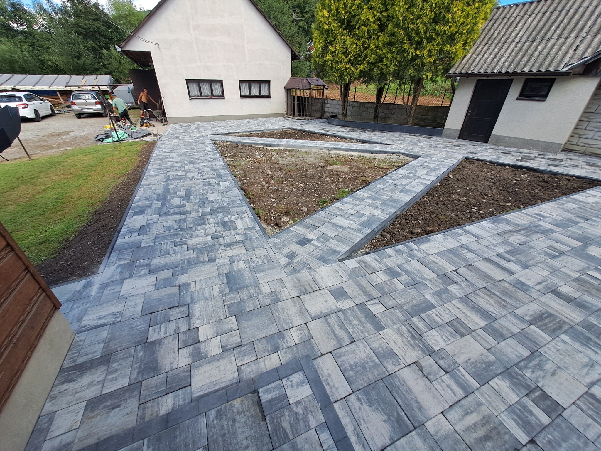Nowoczesna, szara kostka brukowa ułożona w geometryczny wzór na podwórku z fragmentami rabat. W tle budynki mieszkalne i pracujący mężczyźni.