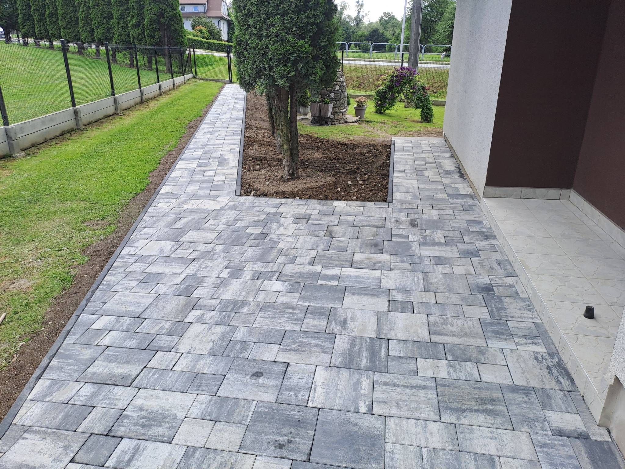 Ułożona kostka brukowa w odcieniach szarości, tworząca ścieżkę wokół drzewa i prowadząca do budynku. Widoczne trawnik, ogrodzenie i elementy zieleni.