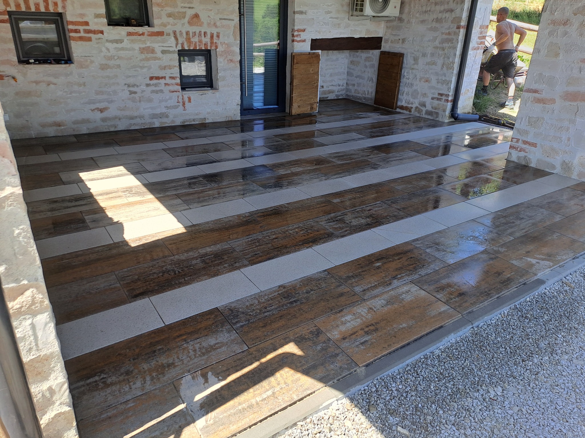 Wykończony taras z płytek imitujących drewno, ułożonych w pasy z szarymi płytkami. Widoczne detale wykończenia i otoczenie tarasu, ściany z cegły i fragment osoby.