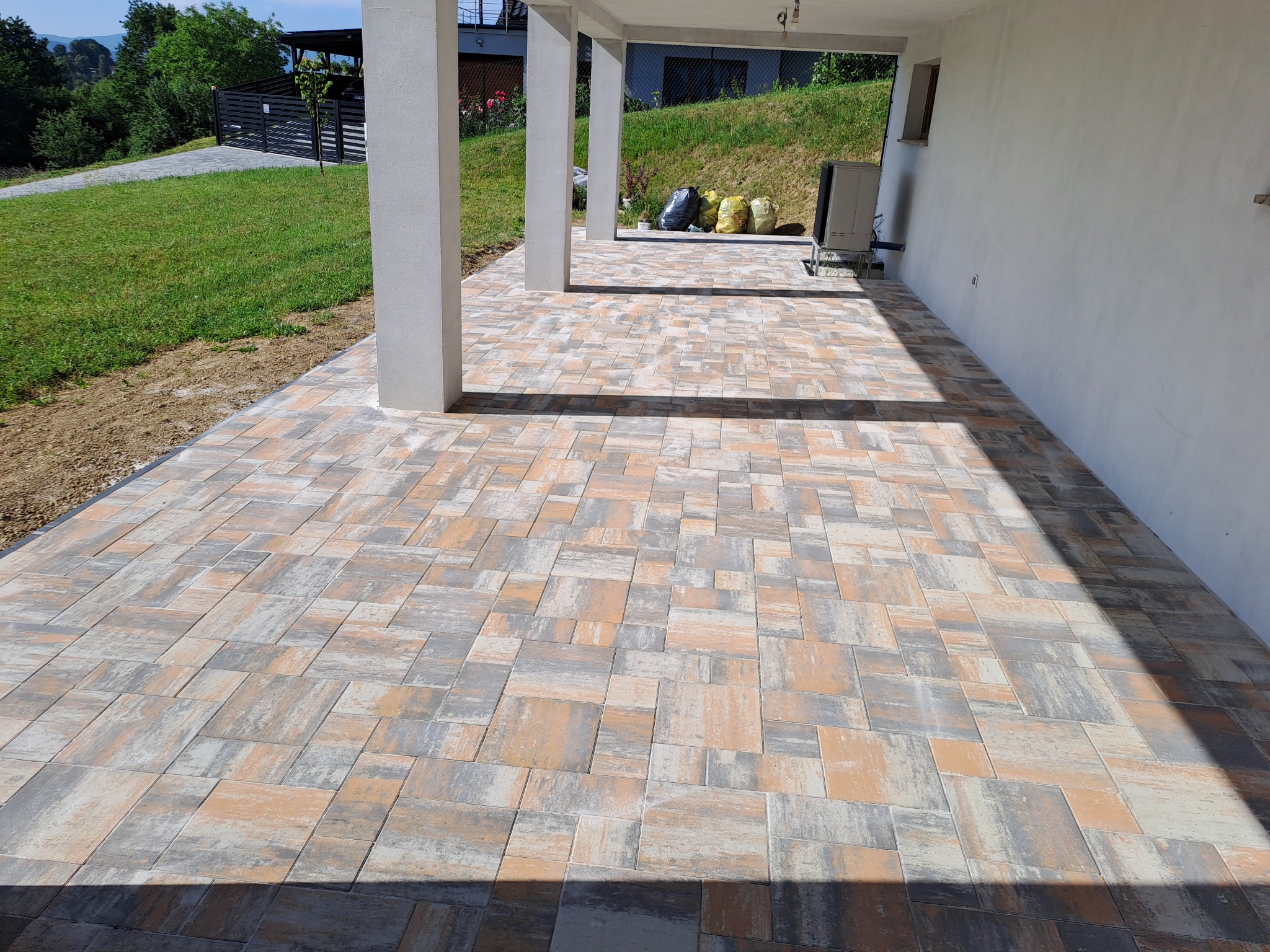 Ułożona kostka brukowa w odcieniach szarości i beżu na zewnątrz budynku z kolumnami. Widoczne worki z materiałami i trawnik w tle.