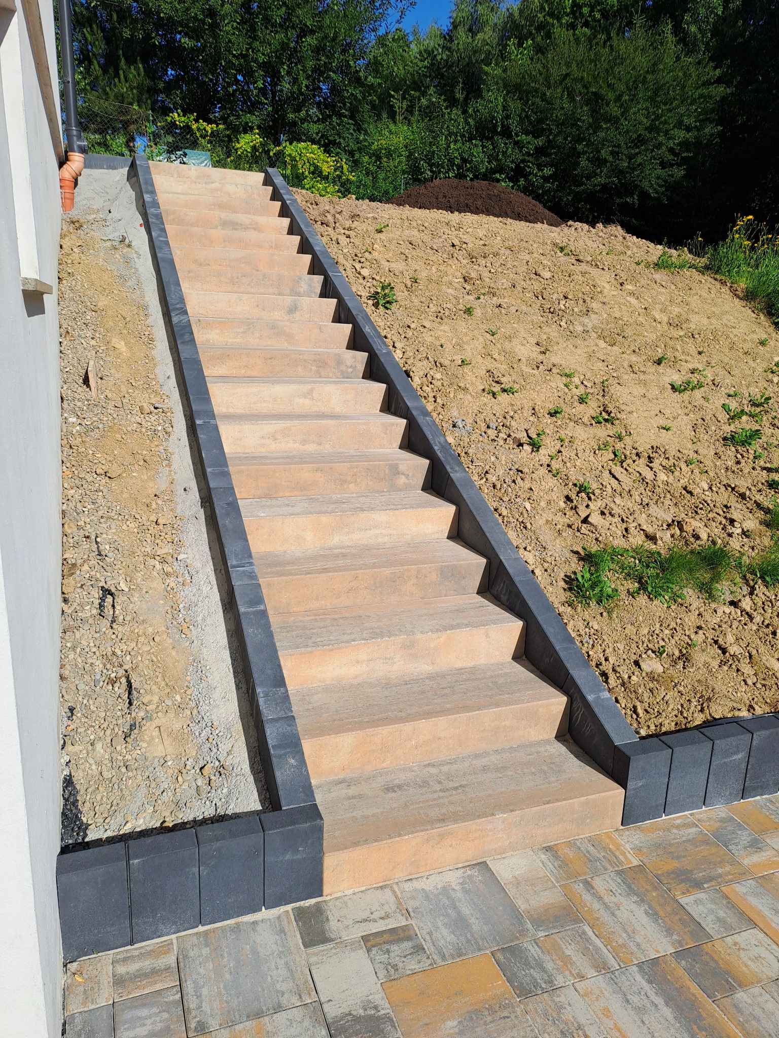 Zewnętrzne schody z kostki brukowej w odcieniach beżu i szarości, prowadzące na wzgórze z ziemią, otoczone zielenią. Widok z dołu, słoneczny dzień.