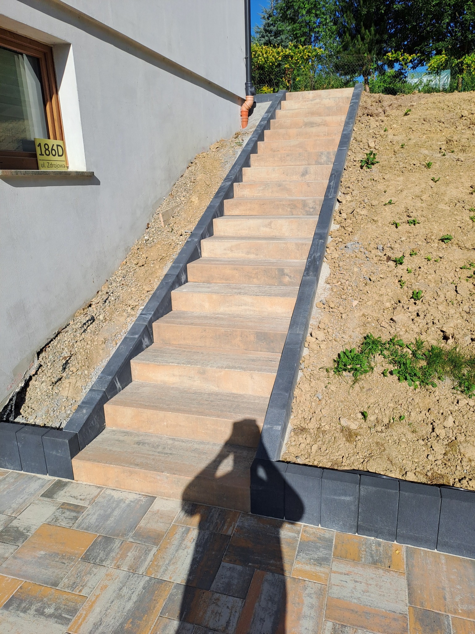 Zewnętrzne schody z kostki brukowej prowadzące na wzniesienie, wykończone ciemnymi obrzeżami. Na dole kostka brukowa ułożona w geometryczny wzór.