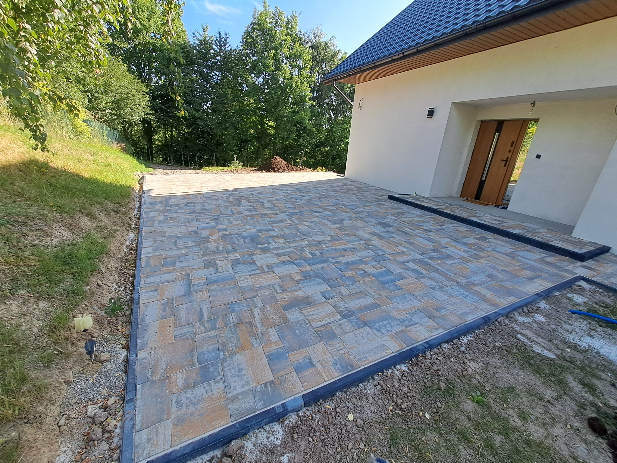 Nowa, kolorowa kostka brukowa ułożona przed nowoczesnym domem z ciemnym dachem. Widoczne obrzeża i fragment trawnika. Słoneczny dzień.
