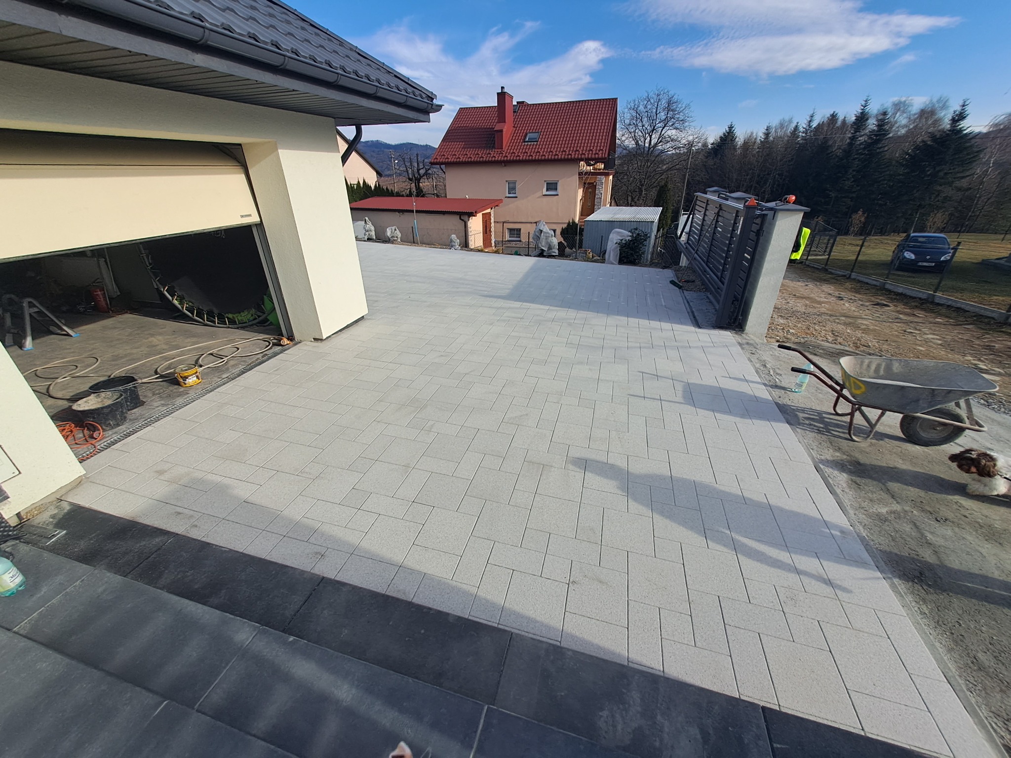 Nowa, jasnoszara kostka brukowa na podjeździe z otwartym garażem i bramą w tle. Widoczne narzędzia i taczka, co sugeruje świeżo zakończone prace.