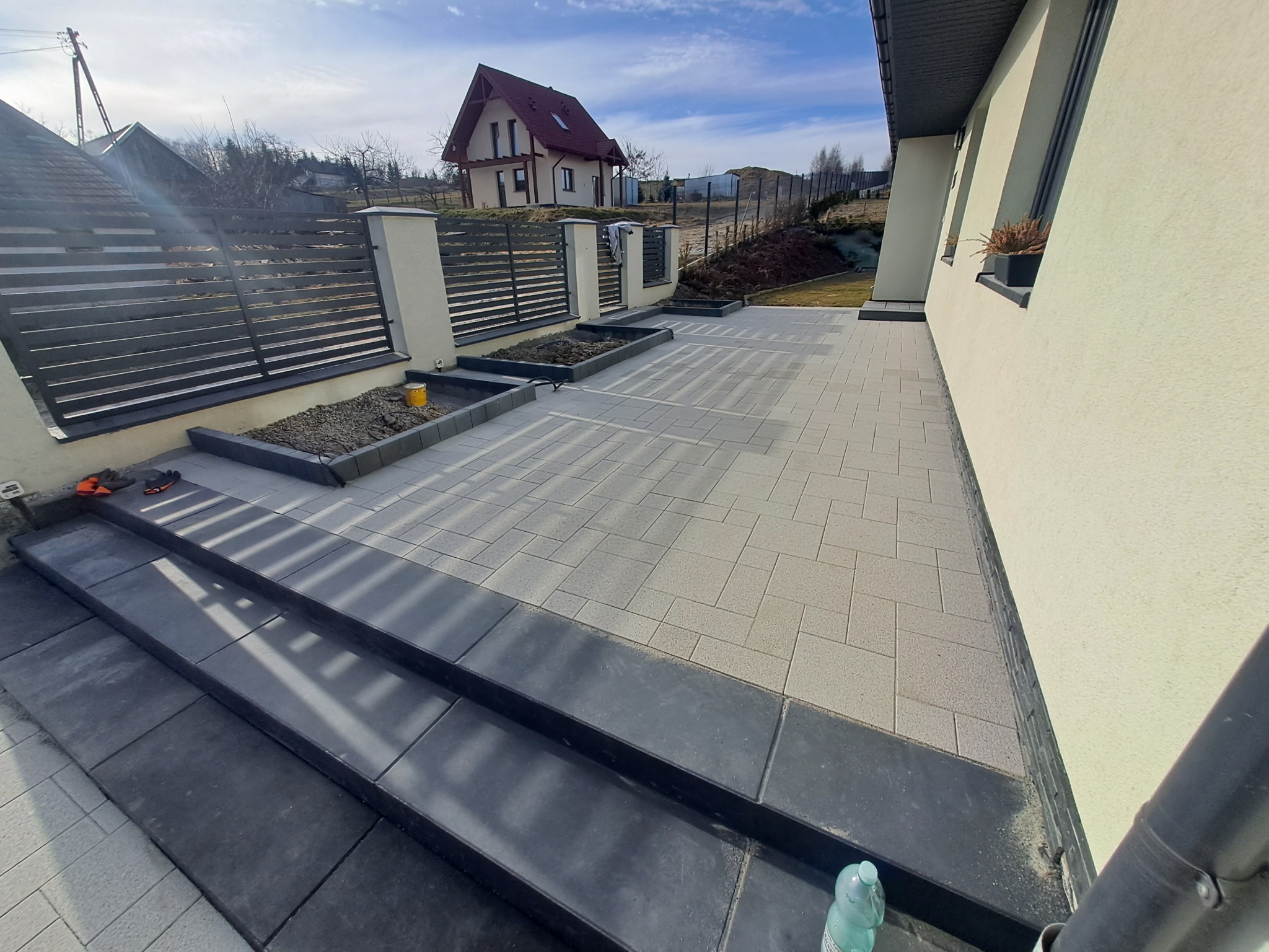 Ułożona kostka brukowa w nowoczesnym stylu, z ciemnymi schodami i rabatami. W tle dom z czerwonym dachem i szare ogrodzenie lamelowe. Słoneczny dzień.