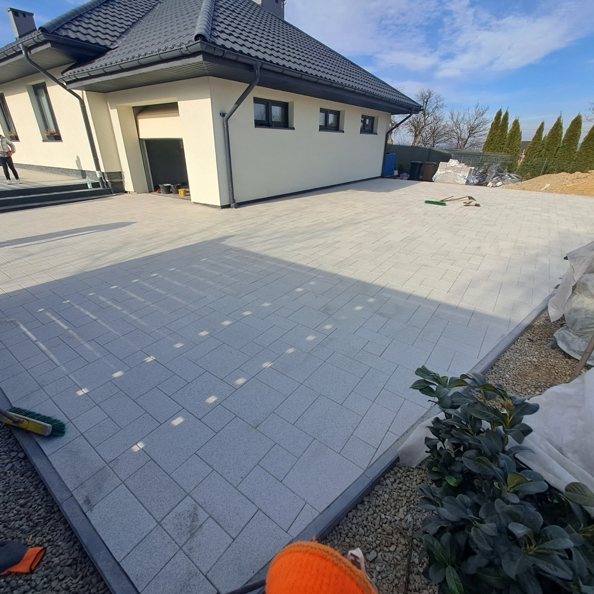Jasnoszara kostka brukowa ułożona na posesji przed domem. Widoczne narzędzia brukarskie. W tle budynek z szarą dachówką i niebieskie niebo.