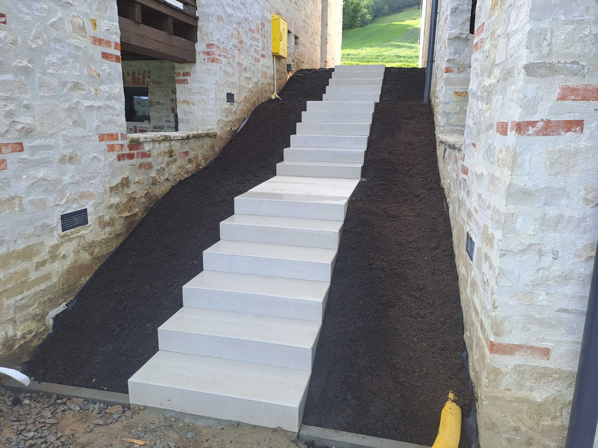 Nowe, jasne schody betonowe prowadzą w górę zbocza między kamiennymi ścianami budynku. Świeża ziemia po bokach schodów. Widok na zielone wzgórze w tle.