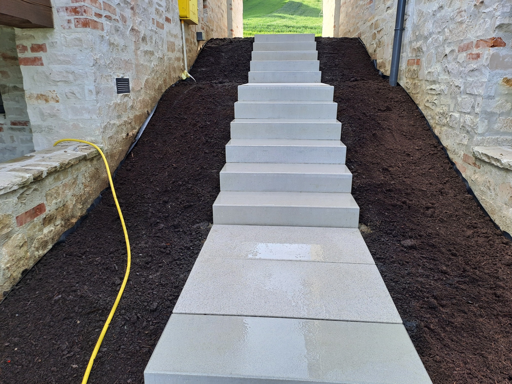 Nowe, jasnoszare schody z kostki brukowej prowadzą w górę, otoczone świeżo nasypaną ziemią i kamiennymi murami. Żółty wąż ogrodowy w lewym dolnym rogu.