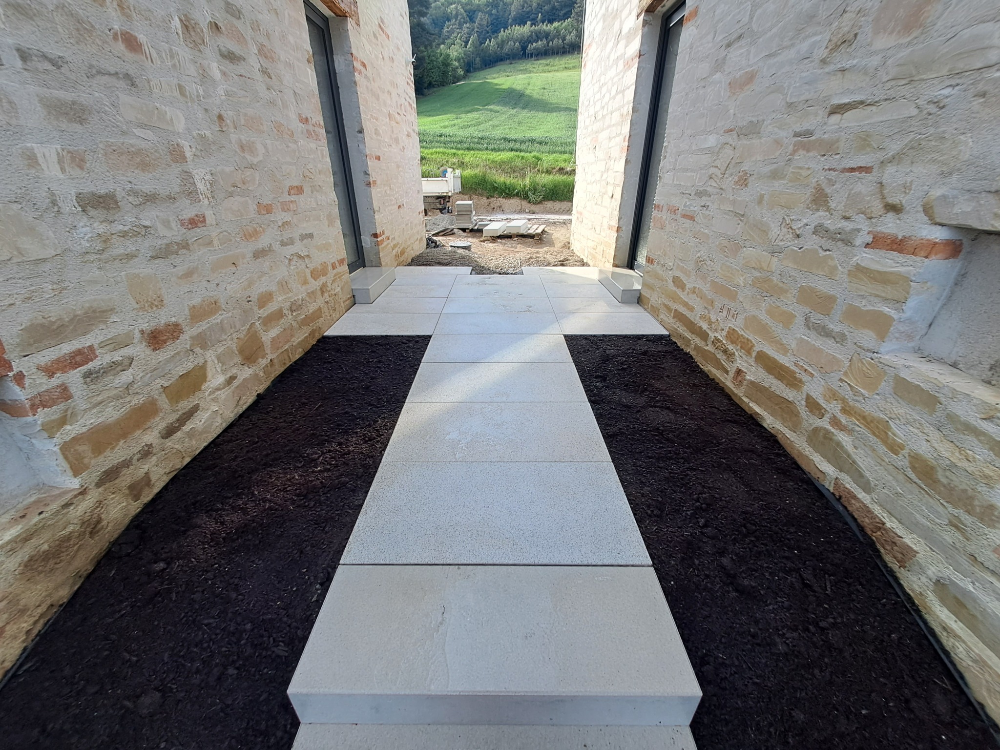Wyłożony jasnymi płytami chodnik prowadzi między kamiennymi ścianami budynku, z widokiem na zielone wzgórze w oddali. Świeżo nasypana ziemia po bokach chodnika.