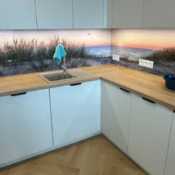 Jasna kuchnia z drewnianym blatem i fototapetą z motywem plaży. Błękitna ściereczka na kranie. Nowoczesne szafki z minimalistycznymi uchwytami. Parkiet w jodełkę na podłodze.