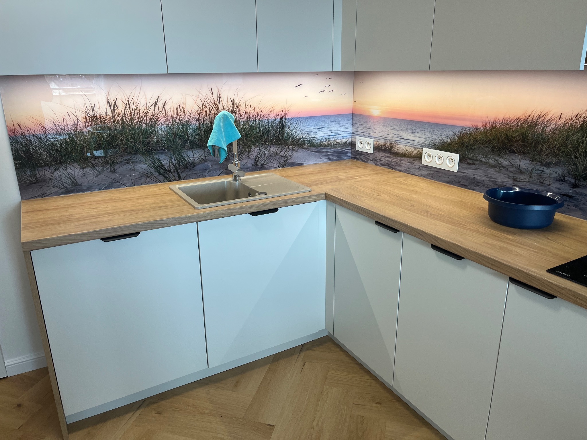 Jasna kuchnia z drewnianym blatem i fototapetą z motywem plaży. Błękitna ściereczka na kranie. Nowoczesne szafki z minimalistycznymi uchwytami. Parkiet w jodełkę na podłodze.