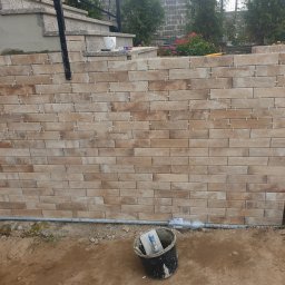 leszek michalski - Układanie beżowych płytek imitujących cegłę na elewacji zewnętrznej, widoczne krzyżyki dystansowe, wiadro z narzędziami na ziemi, fragment schodów i roślinności w tle.