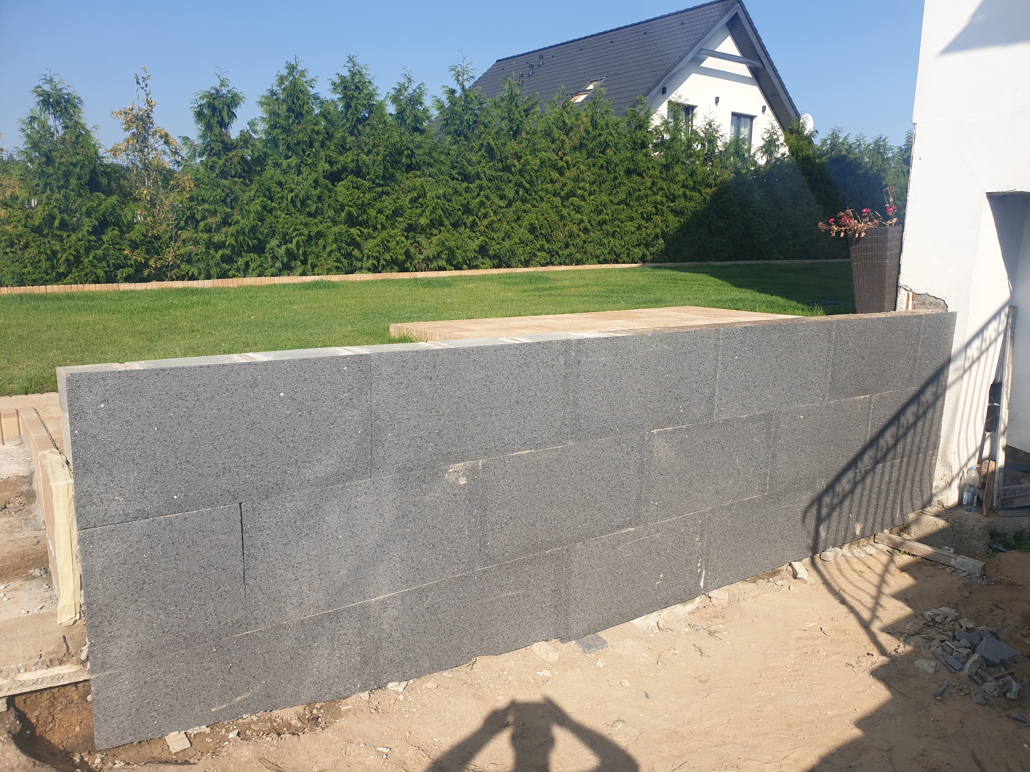 Wznoszenie murku z szarych bloczków betonowych na tle zielonego trawnika, żywopłotu i białego domu z ciemnym dachem, widoczne ślady prac ziemnych i cień osoby robiącej zdjęcie.