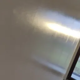 Świeżo nałożona, mokra warstwa na ścianie odbija światło, widoczne ślady po szpachli, okno w tle, podłoga zabezpieczona folią.