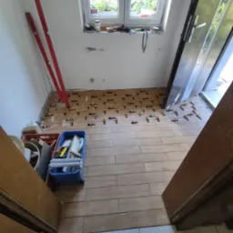 Układanie drewnopodobnych płytek podłogowych z użyciem klipsów poziomujących, widoczne narzędzia i poziomica oparta o ścianę.