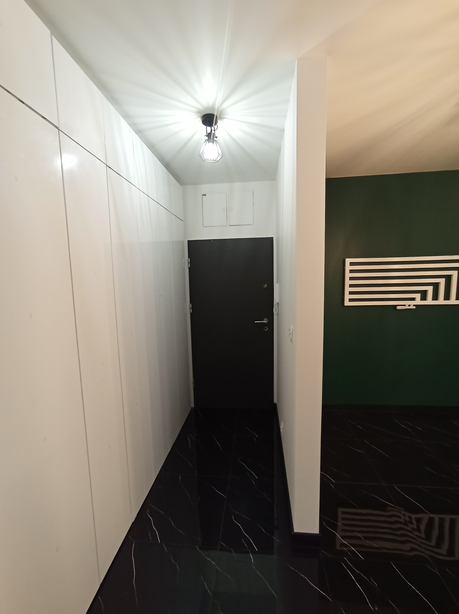 Nowoczesny korytarz z czarną, marmurową podłogą, białymi szafami w zabudowie i ciemnozieloną ścianą z dekoracyjną, białą ozdobą.