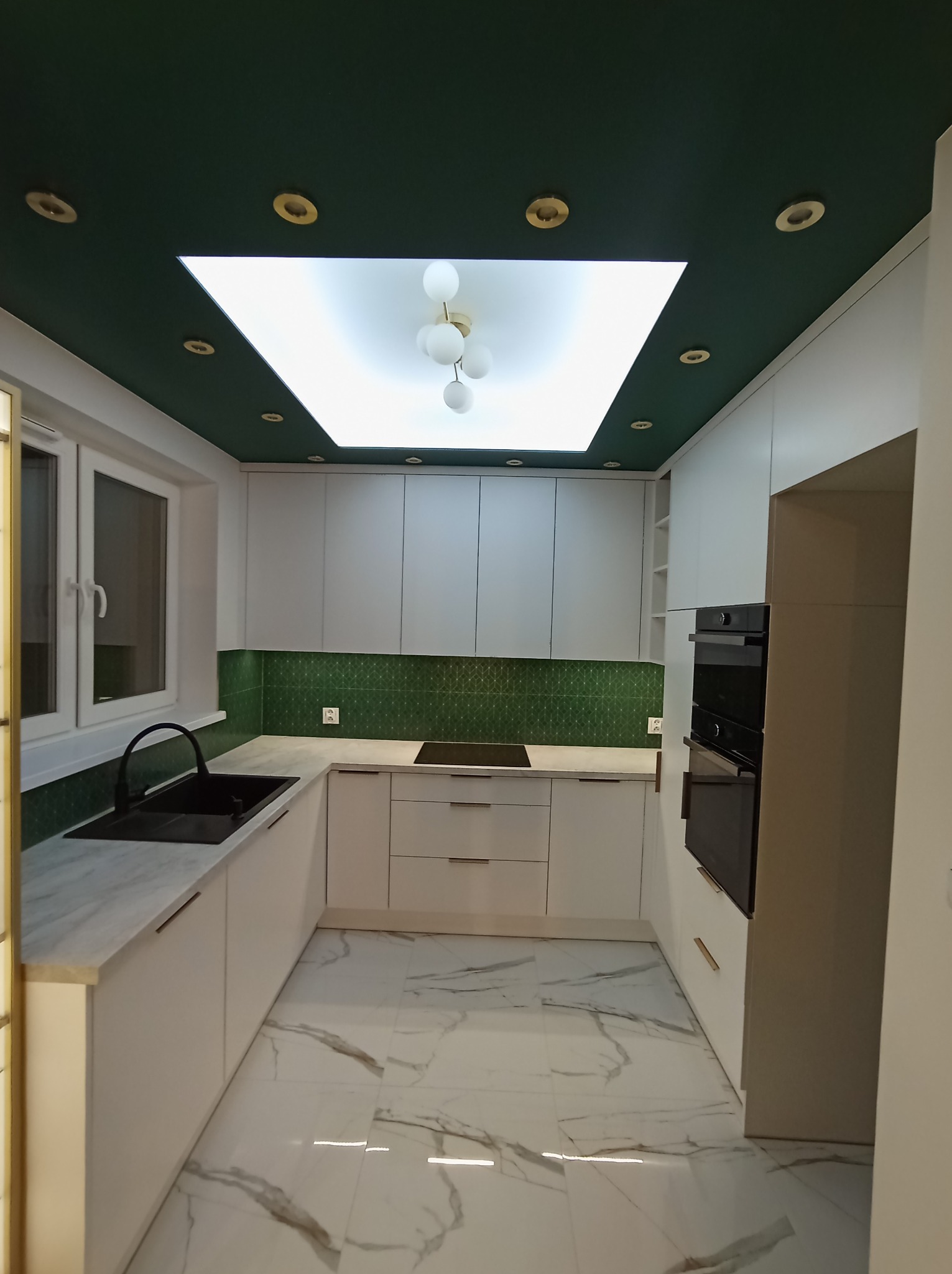 Nowoczesna kuchnia w bieli z zielonym akcentem na suficie i nad blatem, białe szafki z marmurowymi blatami i podłogą, czarna armatura i sprzęt AGD, oświetlenie sufitowe.