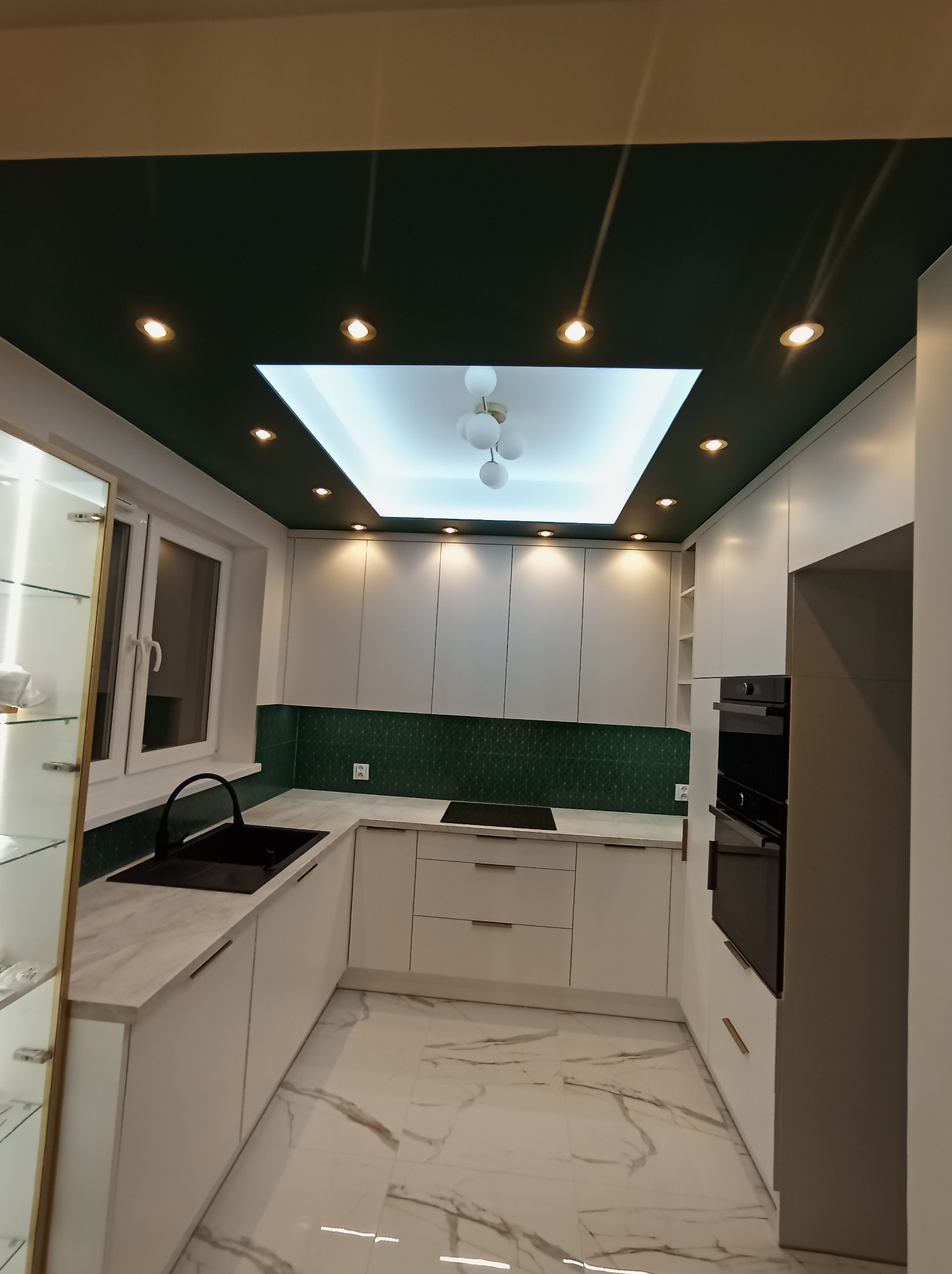Nowoczesna kuchnia z białymi szafkami, marmurową podłogą i blatem, zielonymi płytkami nad blatem oraz ciemnozielonym sufitem z oświetleniem punktowym i centralnym.